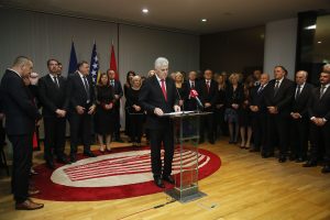zajedničko djelovanje u korist bolje budućnosti Dragan Čović stoji za govornicom staklenom iza njega mnogo ljudi u sali crveno bijeli ćilim tamno odijelo bijela košulja crvena kravata sneđi parket zastave