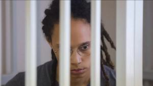 Brittney Griner Afroamerikanka sa dugom kosom crnom naočale siva majica vidi se do ramena iza bijelih rešetaka zatvorskih iza sivi zid spuštenog pogleda djeluje zabrinuto