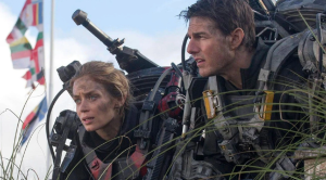 Emily Blunt i Tom Cruise u sceni iz filma Edge of Tommorow vide se do struka gledaju negdje u daljinu poluprofil na njima kostimi puni žica željeza i oružja prljava lica dan maslonastosiva odijela sa bordo detaljima iza poderana zastava nejasno čija i par grančica