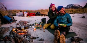 Norvežani friluftsliv sjede muškarac i žena vani pored zapaljene vatre hrana ispred njih i piće u drvenim čašama kao za vino oni obučeni u jakne i čizme okolo snijeg zalazak sunca