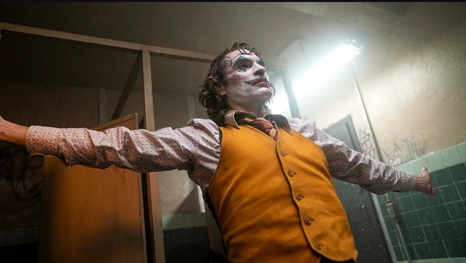 sa snimanja nastavka Jokera Joaquin Phoenix u narandžastom prsluku roze košulji raširenih ruku na licu jaka šminka kao joker sa bijelim puderom crnim ustima u prostoriji kroz prozor probija dnevna svjetlost iza drvena vrata