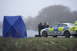 Nezapamćena tragedija u Velikoj Britaniji policijski automobil bijele boje piše policija sa plavobijelim kockama pored tri policajca vide se s leđa lijevo od njih plavi šator ispred jezero u daljini drveće u magli zimski dan pored trava napola zaleđena