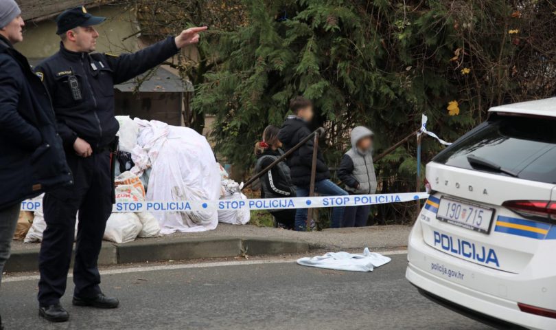 klasična sačekuša u Zagrebu policajac na ulici u uniformi pokazuje lijevom rukom na nešto u daljini drugom policajcu kapa na glavi ispred policijsko auto bijelo lijevo bijele vreće pored stoji troje djece u jaknama lica su im zamagljena bor oblačan dan