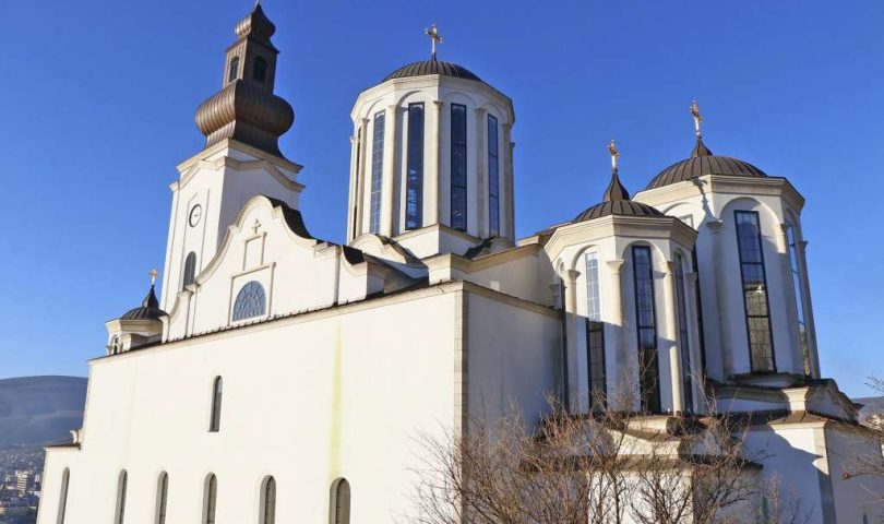 Pokradena Saborna crkva Saborna crkva u Mostaru bijelo veliko zdanje sa tri kule i jednim zvonikom na prednjem zidu šest prozora drvo uz desnu stranu crkva plavo nebo bez oblaka dan