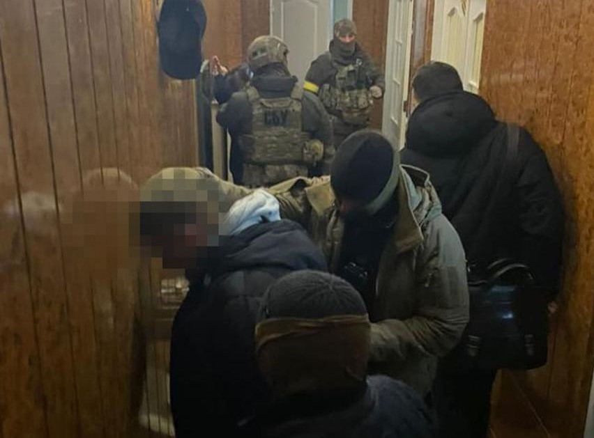 uhapsili bračni par ukrajinska vojska pet njih sa fantomkama na glavama u hodniku smeđi drveni zid drže muškarca kojem je zamagljeno lice prislonjenog uz zid okrenut ka zidu crna jakna bijela kapuljača na zidu nešto crno okačeno na dnu hodnika bijela vrata uzak prostor