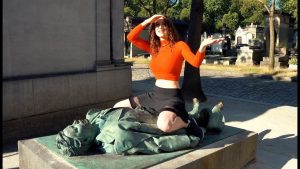 Najperverzniji grob žena u narandžastoj majici i crnom miniću sjedi na skulpturi na grobu rukom zaklanja sunce na očima duga crna kosa okolo grobovi i drveće dan