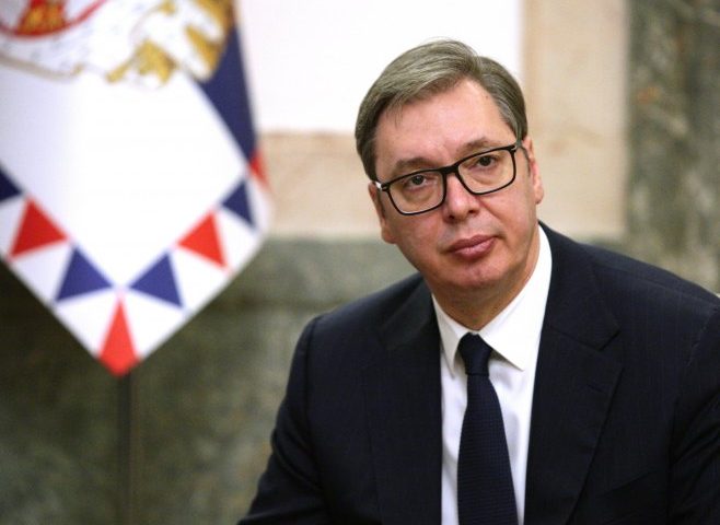dati državu Srbiju na tacni Aleksandarc vučić crno odijelo bijela košulja tamna kravata naočale sjedi u uredu iza njega sivo i zastava iza